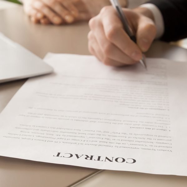 Close up image of paper contract document lying at work desk with laptop, businessman putting his signature under text of financial agreement with ballpoint pen in presence of female business partner